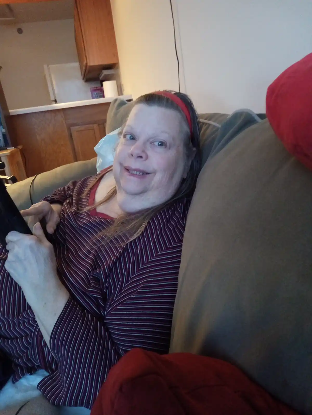 A woman with a red headband and striped shirt sits on a couch, holding a remote, with a wide-eyed, slightly amused…
