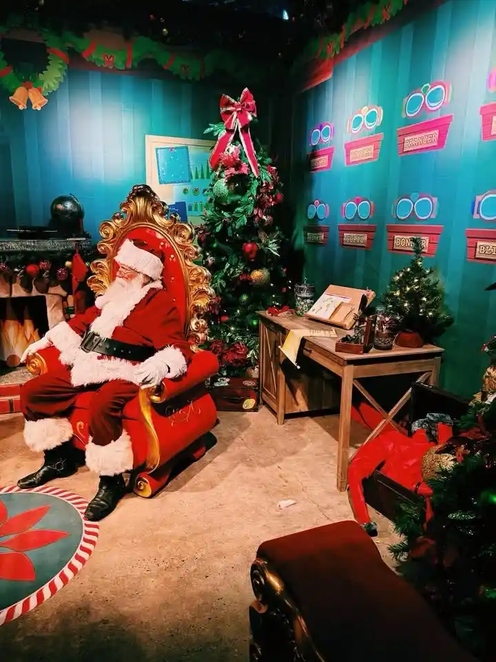 A person dressed as Santa Claus is slumped over in a large red throne chair in a festive, decorated room with Christmas…
