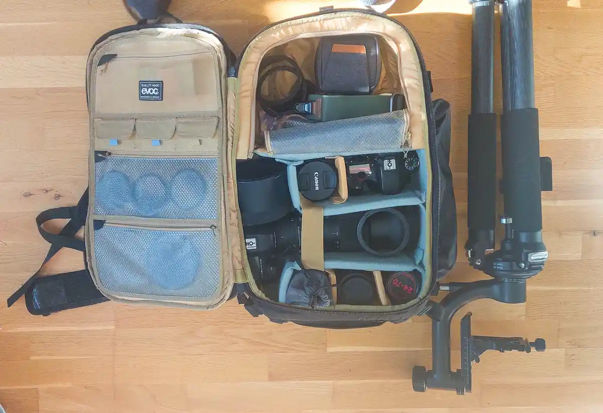 An open, beige Evoc camera backpack lies on a wooden floor, packed with multiple lenses, a Canon DSLR camera, and other…
