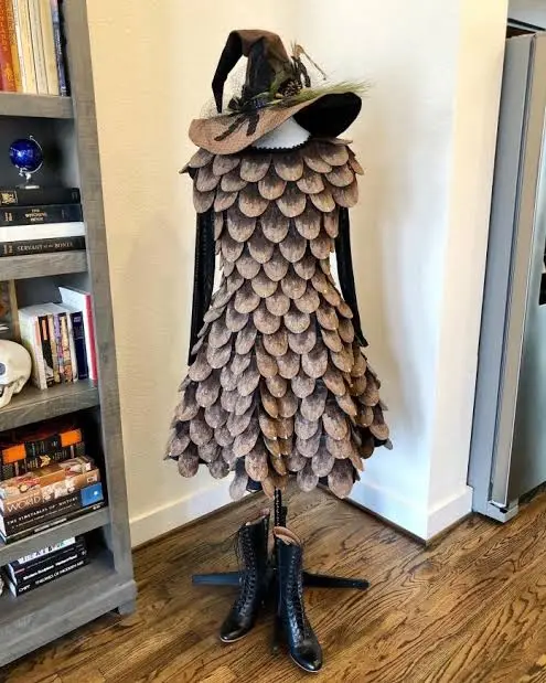 A mannequin in a room stands wearing a full costume made entirely of pinecones, including a hat and boots placed at its…