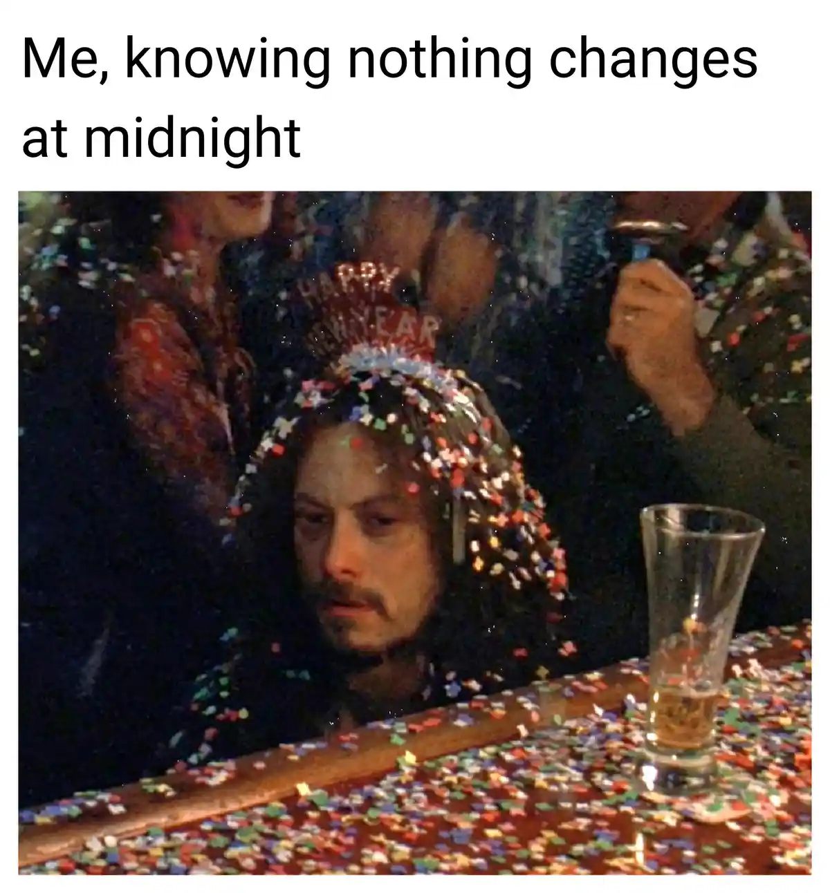 A man with a weary, resigned expression sits at a table covered in colorful confetti, with a nearly empty glass beside him…