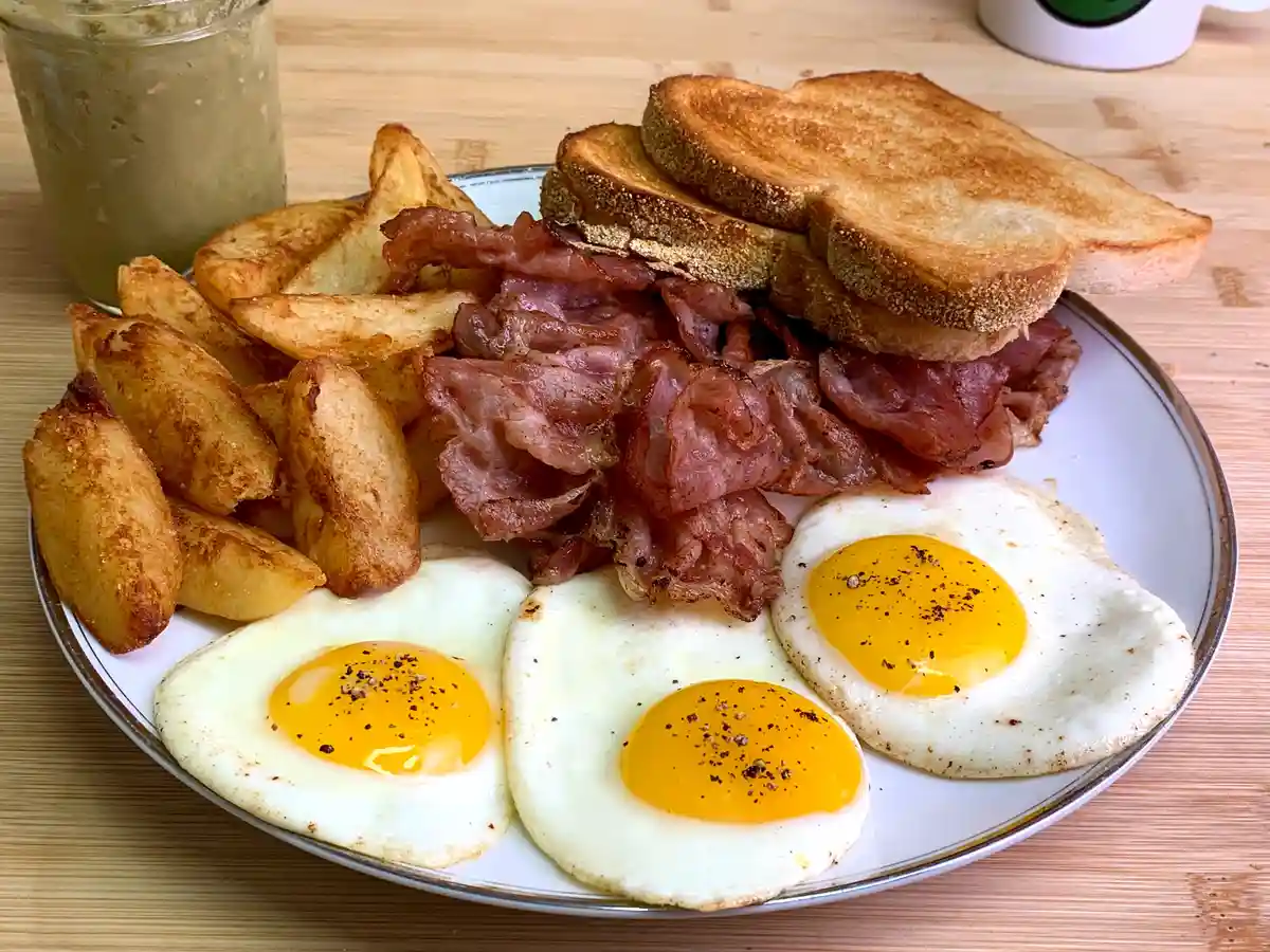 A plate of breakfast food including three fried eggs, crispy bacon, golden-brown potato wedges, and several slices of…