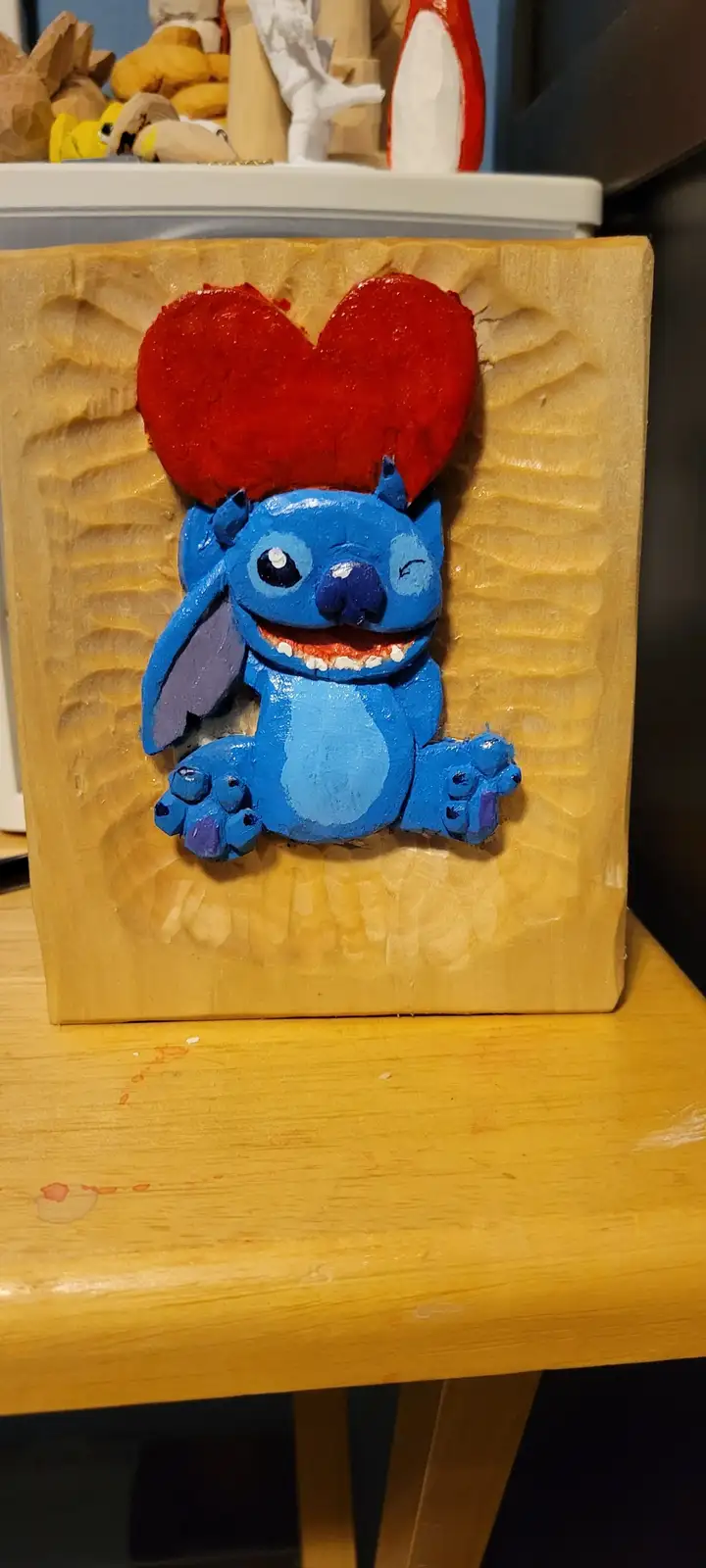 A hand-carved wooden relief depicts the blue Disney character Stitch with a large red heart above his head, set against a…