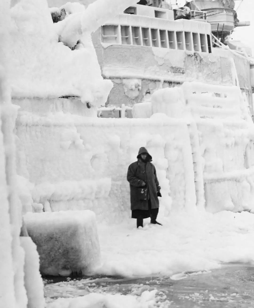 A black-and-white historical photo shows a person in a heavy winter coat standing in front of a massive ship completely…