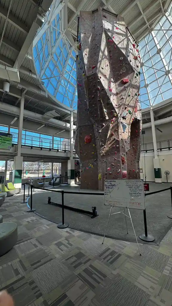 A tall indoor climbing wall with colorful handholds is surrounded by a black stanchion rope barrier inside a large, modern…