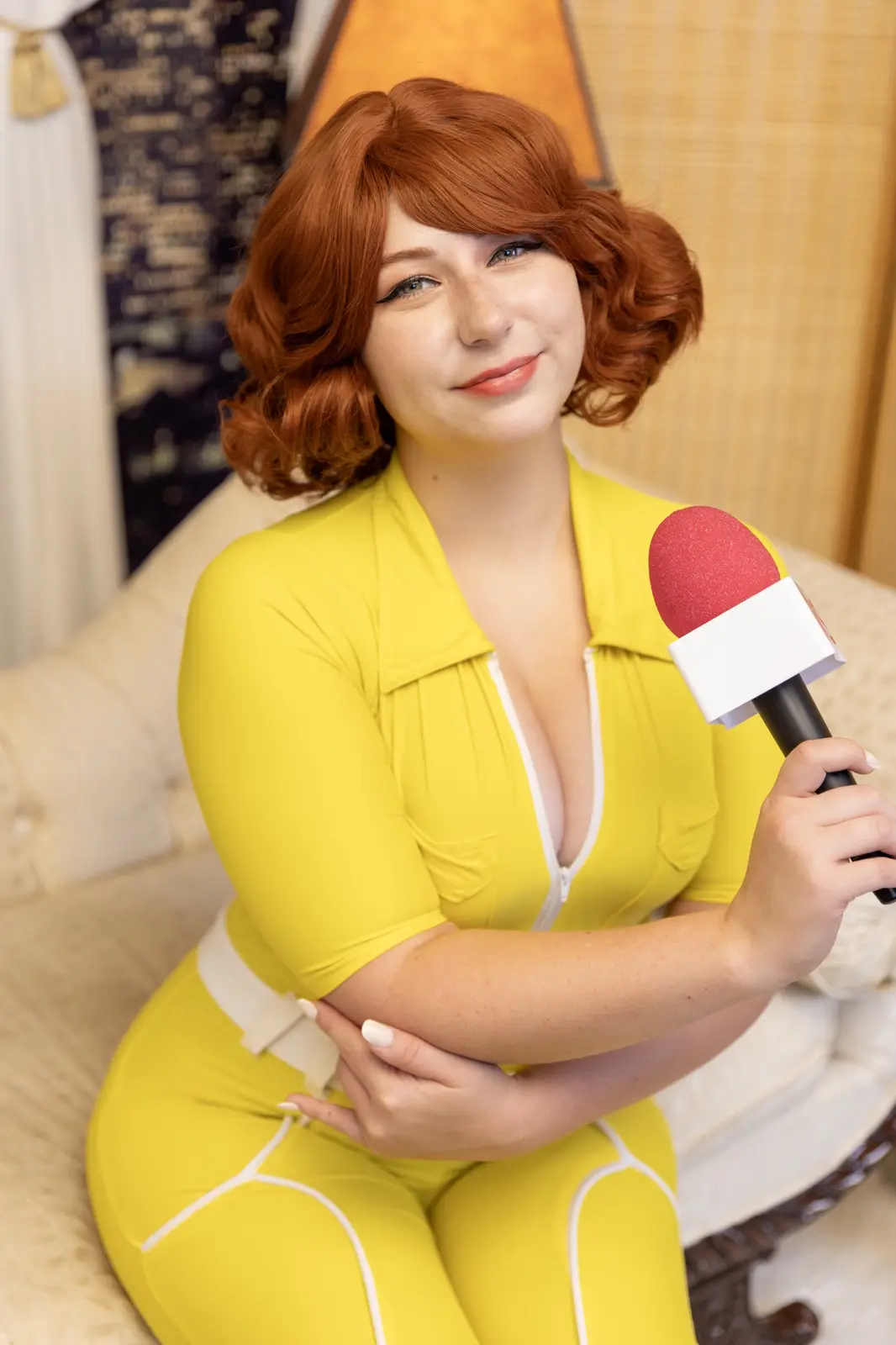 A person in a yellow cosplay outfit with red hair holds a microphone while sitting on a couch, smiling. The overlaid text…