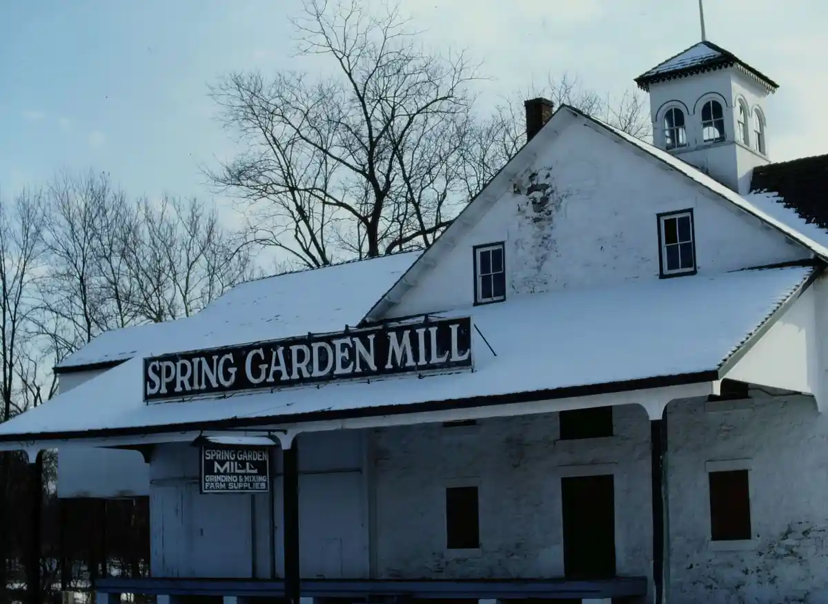 A snowy, white-painted building with a sign reading 'SPRING GARDEN MILL' and a smaller sign below stating 'GRINDING & MIXING…