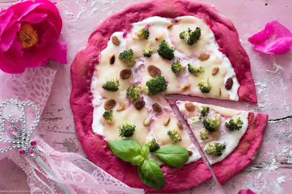 a pink-colored pizza with a pink crust, topped with white sauce, broccoli florets, raisins, and pine nuts, sits on a pink…