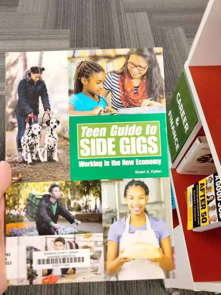 A library book titled 'Teen Guide to SIDE GIGS Working in the New Economy' is held up, showing images of teens doing various…