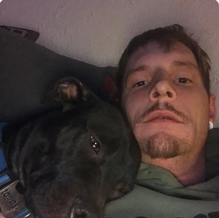 A man with a goatee and ear piercings lies in bed next to a black dog, both looking at the camera with a tired expression…