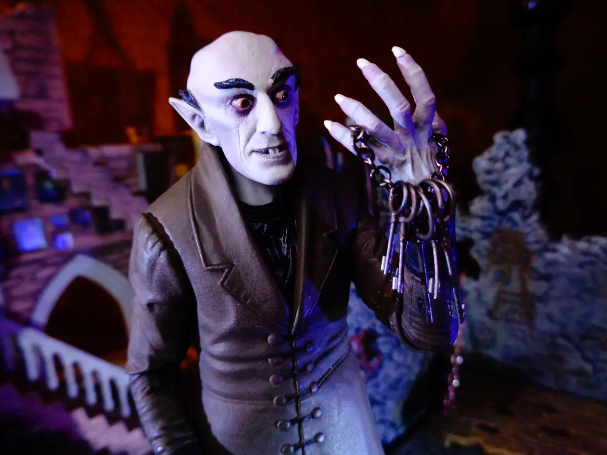 A figurine of a bald, pale-skinned vampire with pointed ears and fangs, wearing a brown vest over a black shirt, holds a…