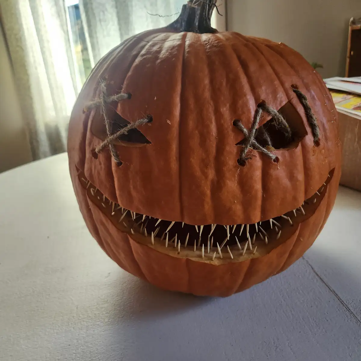 A close-up photo of a carved orange pumpkin with triangular eye holes stitched shut with twine and a wide, jagged mouth…