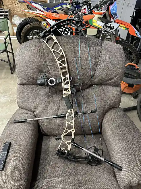 A modern compound bow with a tan and black frame, blue strings, and a mounted sight is propped upright on a brown fabric…
