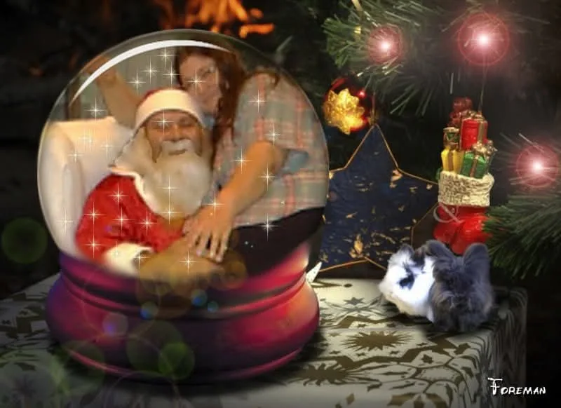 A snow globe contains a photo of Santa Claus sitting on a couch with a woman leaning over him, both smiling. The globe sits…