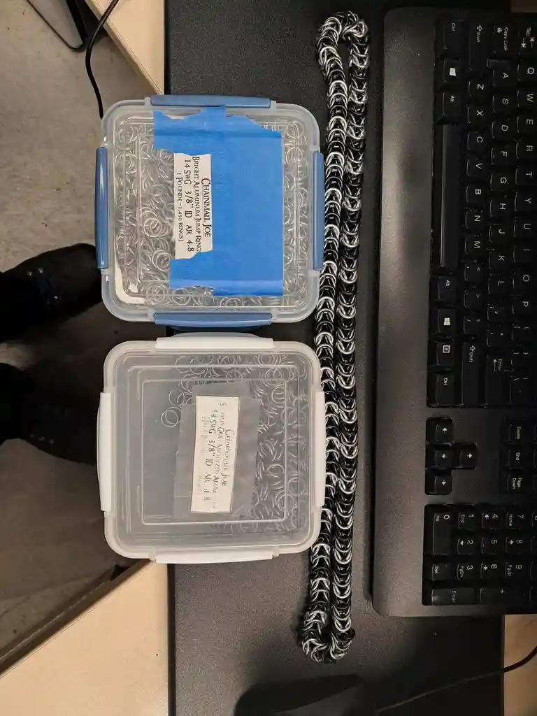Two clear plastic containers filled with small metal jump rings sit on a desk next to a black keyboard, with a long…