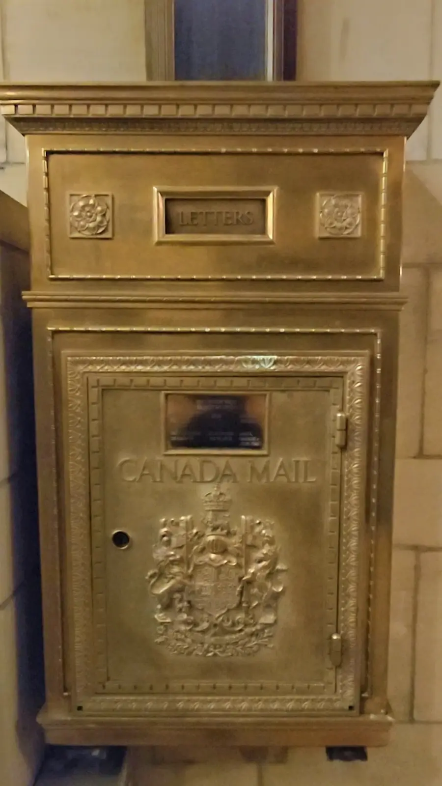 A close-up of an ornate, golden Canada Mail postbox with 'LETTERS' engraved above the slot and the Canadian coat of arms…