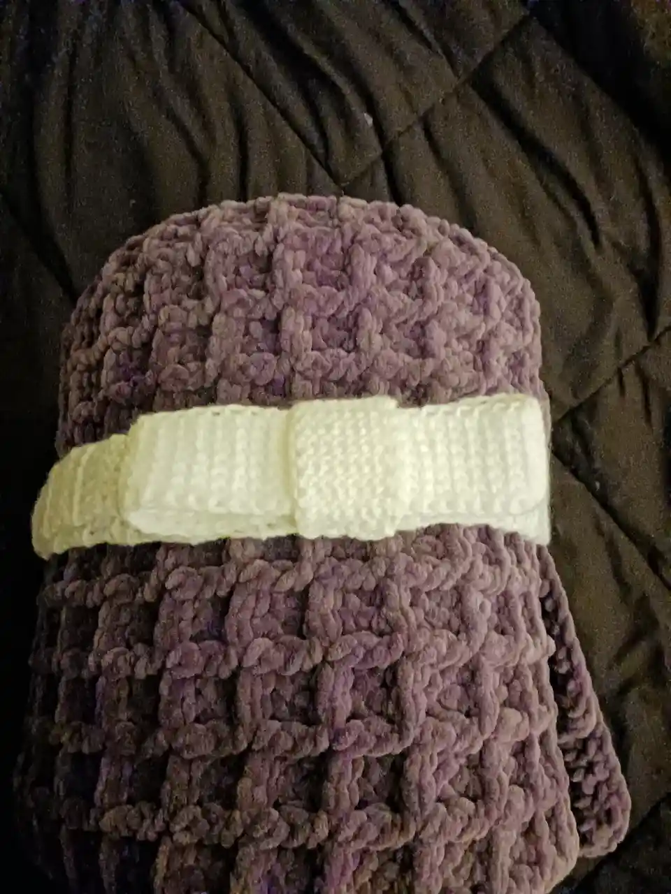 A close-up photo shows a thick, textured, purple crocheted item, possibly a hat or blanket, with a wide, cream-colored…