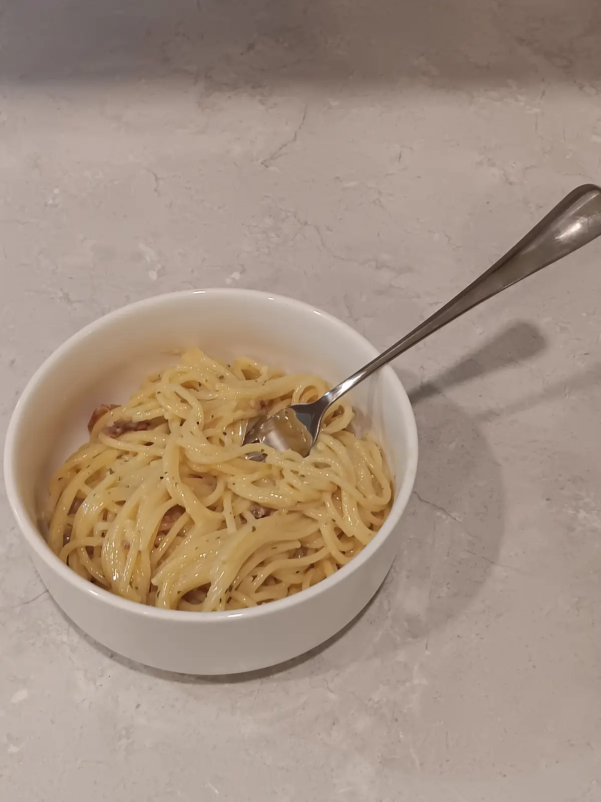 A white bowl filled with plain spaghetti, lightly coated in sauce and garnished with black pepper and herbs, sits on a light…