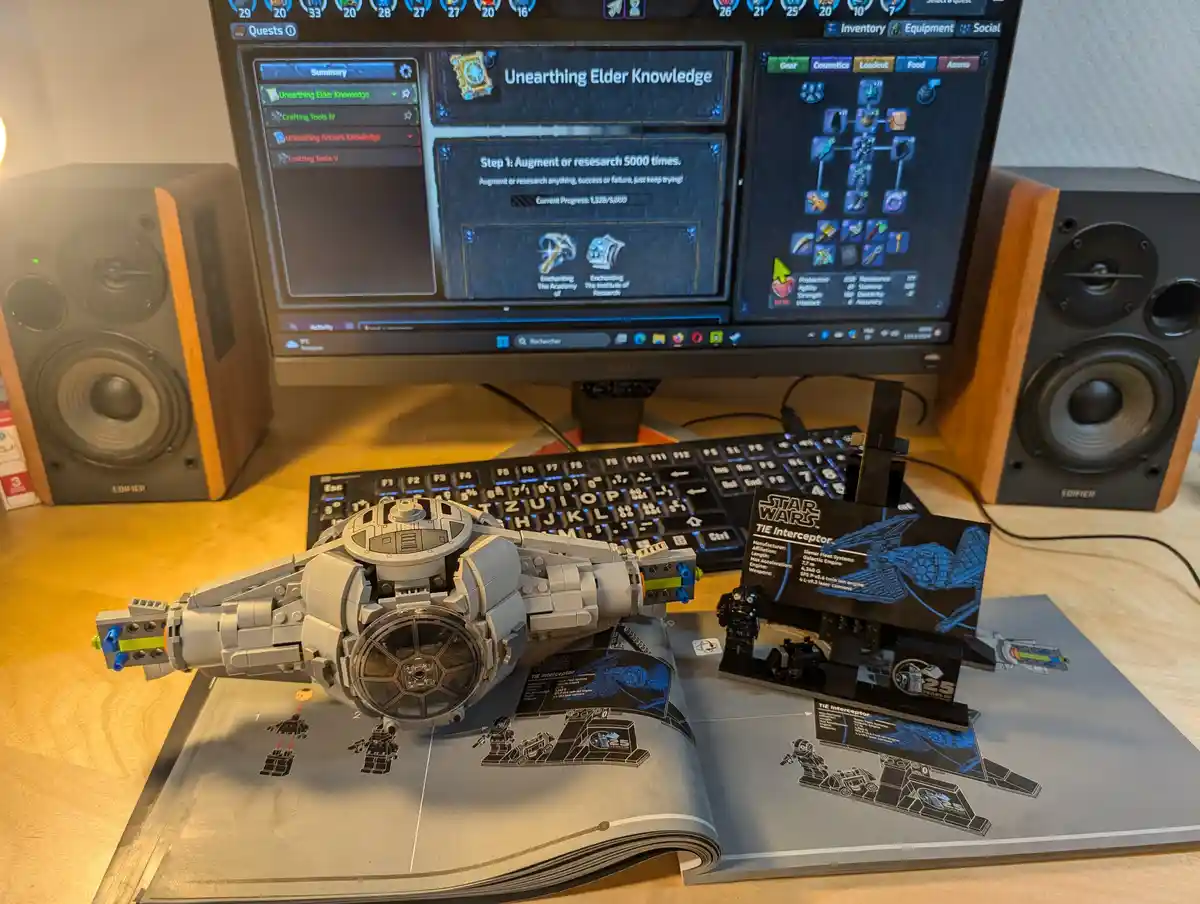 A completed LEGO Star Wars TIE Interceptor model sits on its instruction manual on a desk, with a computer monitor in the…