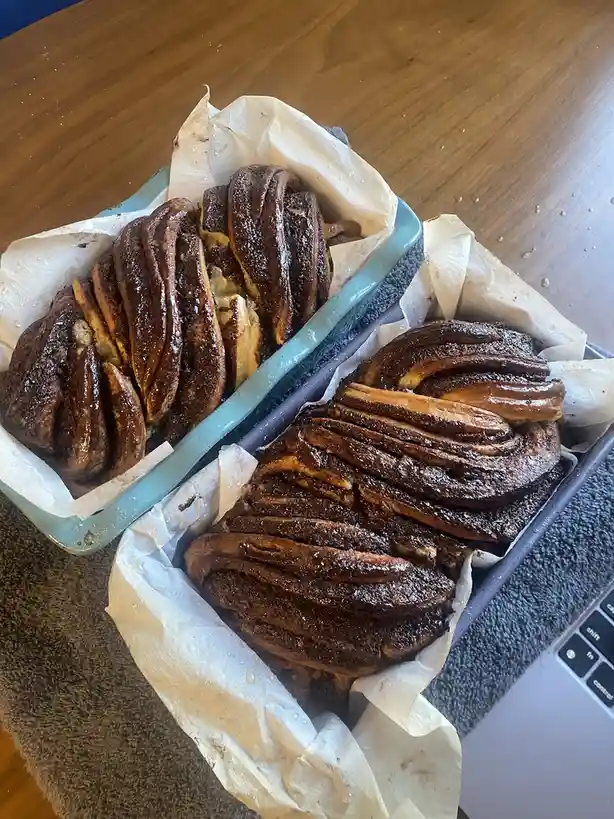 Two rectangular loaf pans sit on a towel, each containing a large, darkly browned, caramelized bread loaf with a glossy…