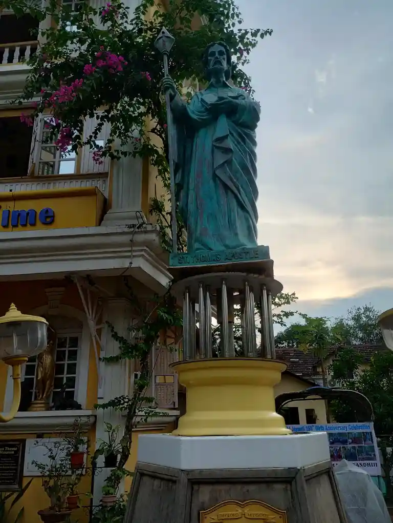A weathered green statue of St. Thomas Apostle stands on a yellow pedestal in front of a yellow building with white trim and…