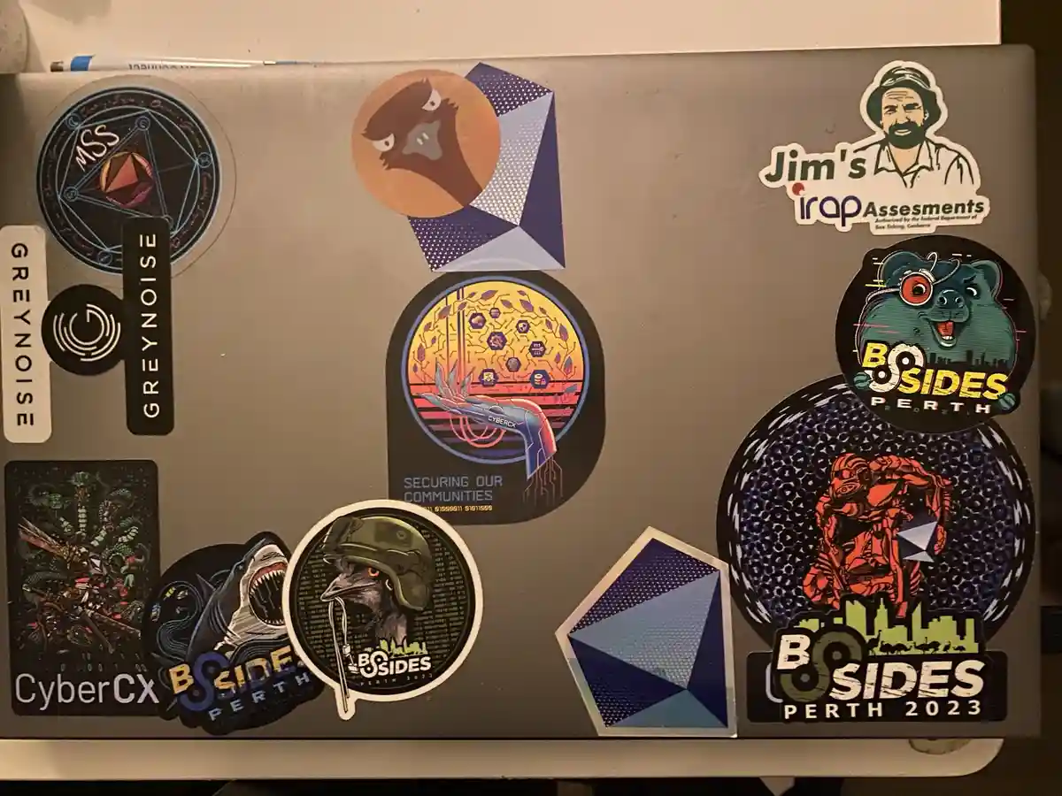 A laptop lid covered in numerous stickers, including multiple 'BSides Perth' event stickers, 'GreyNoise' branding, a…