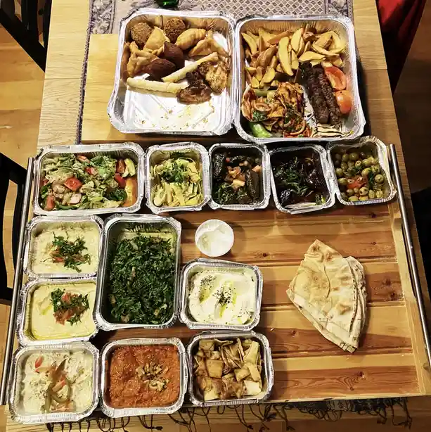 An overhead photo shows a wooden table covered with numerous aluminum trays filled with a wide variety of Middle Eastern and…