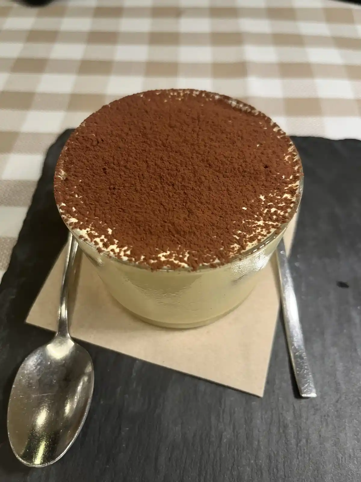 A close-up photo shows a glass cup filled with creamy tiramisu, generously dusted with cocoa powder, resting on a brown…