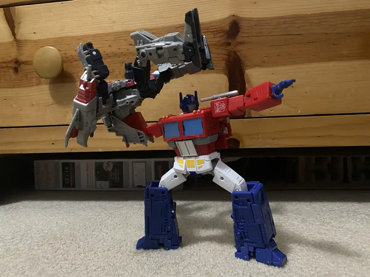 A red, blue, and white Optimus Prime action figure stands on a beige carpet, holding up a smaller grey and red Transformer…