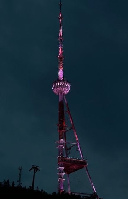 A tall, slender tower with a spherical middle section is illuminated with pink and purple lights against a dark night sky…