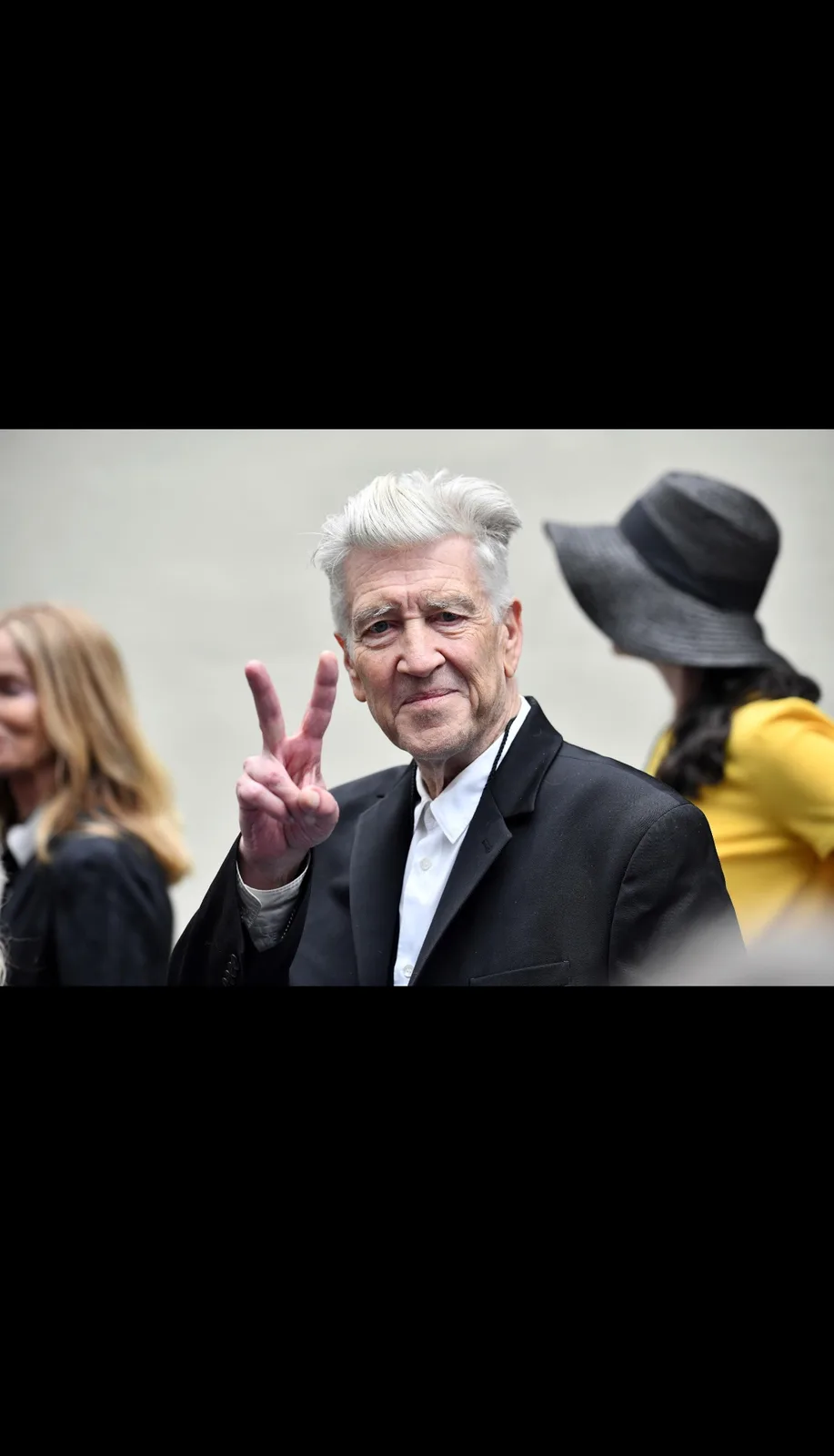 An older man with white hair, wearing a black suit and white shirt, makes a peace sign with his right hand while looking…