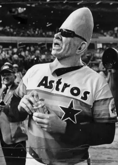A black-and-white photo of a man wearing a Houston Astros baseball jersey and a large, cone-shaped alien headpiece, holding…