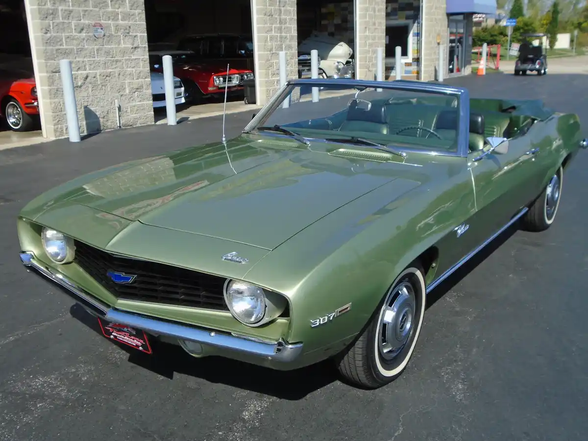 A shiny, olive-green 1967 Chevrolet Camaro convertible is parked outside a car dealership with other classic cars visible in…