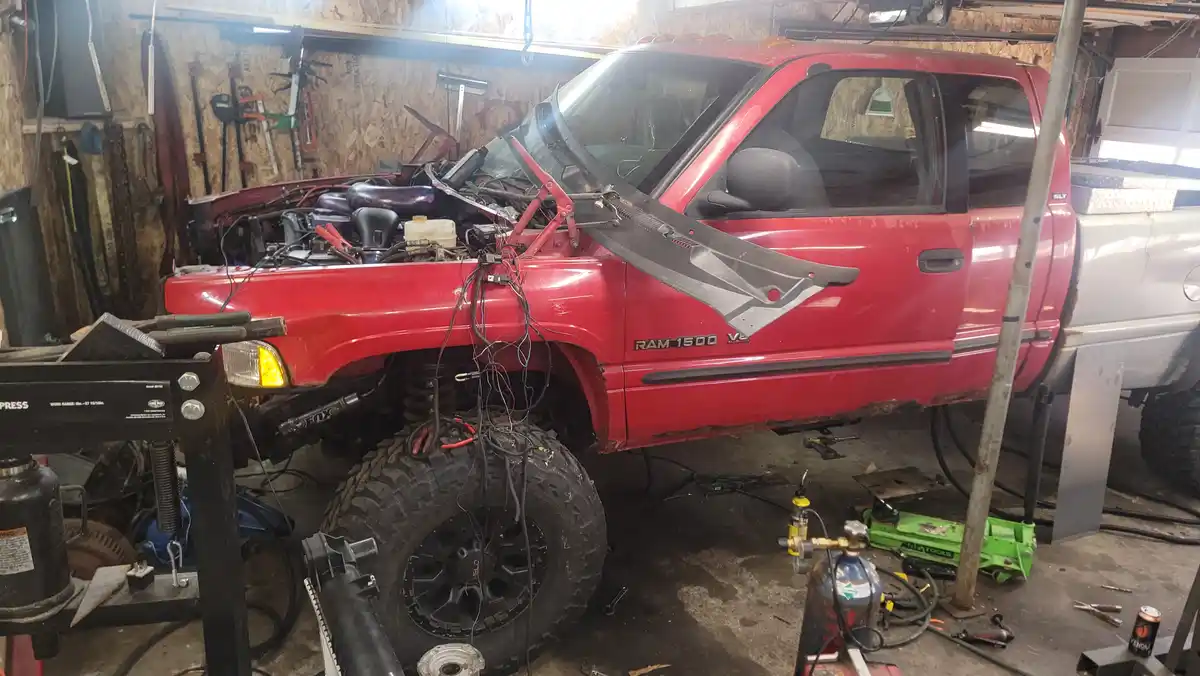 A red Dodge Ram 1500 pickup truck is shown in a cluttered garage with its hood open, engine exposed, and front end heavily…