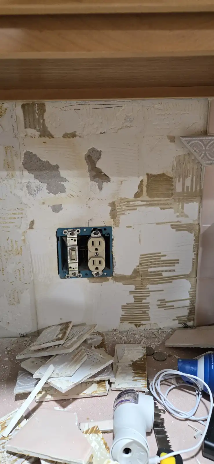 A close-up photo shows a wall under renovation with peeling, damaged wallpaper and broken tiles on the floor. A blue…