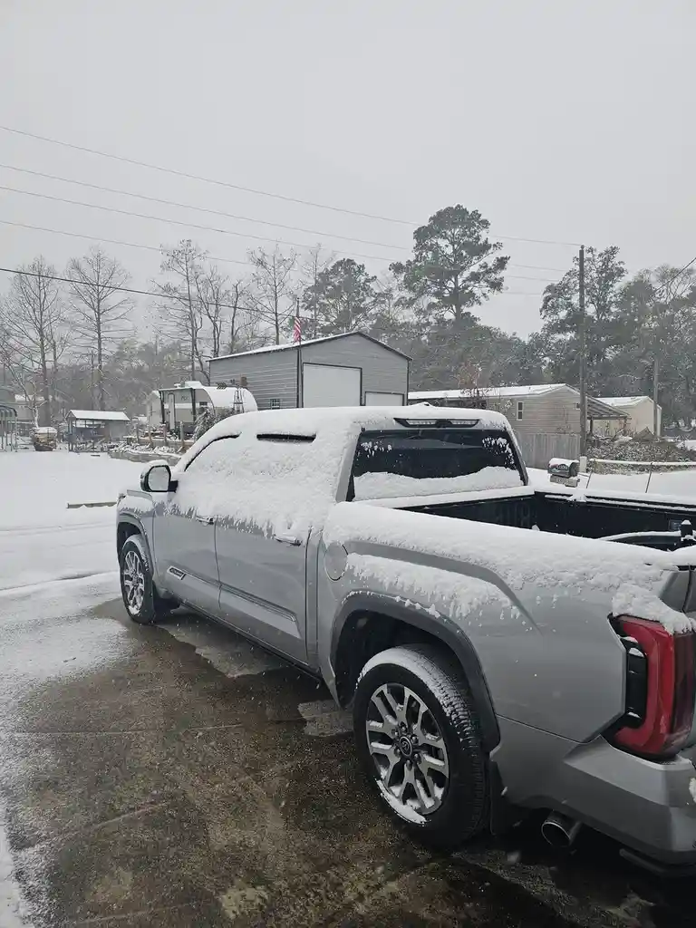 A silver pickup truck is parked in a snowy residential area, its entire roof, bed, and upper body covered in a thick layer…
