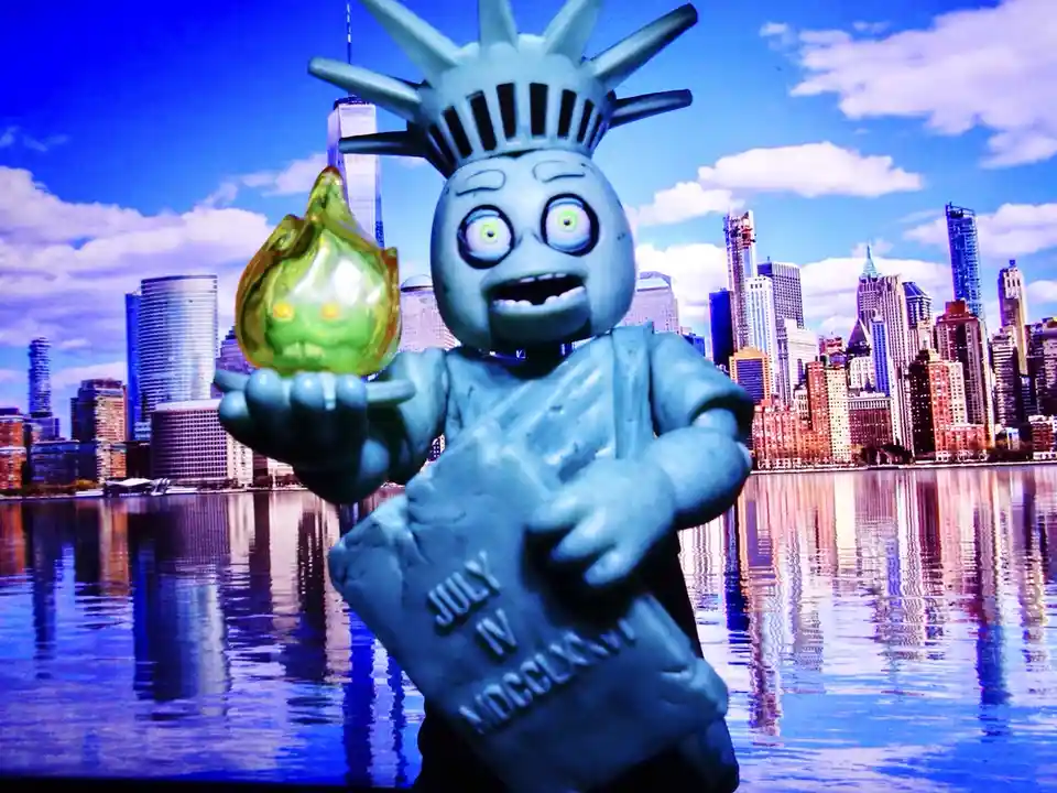 A small, blue, cartoonish Statue of Liberty figurine with wide, green eyes and a surprised expression stands in front of a…