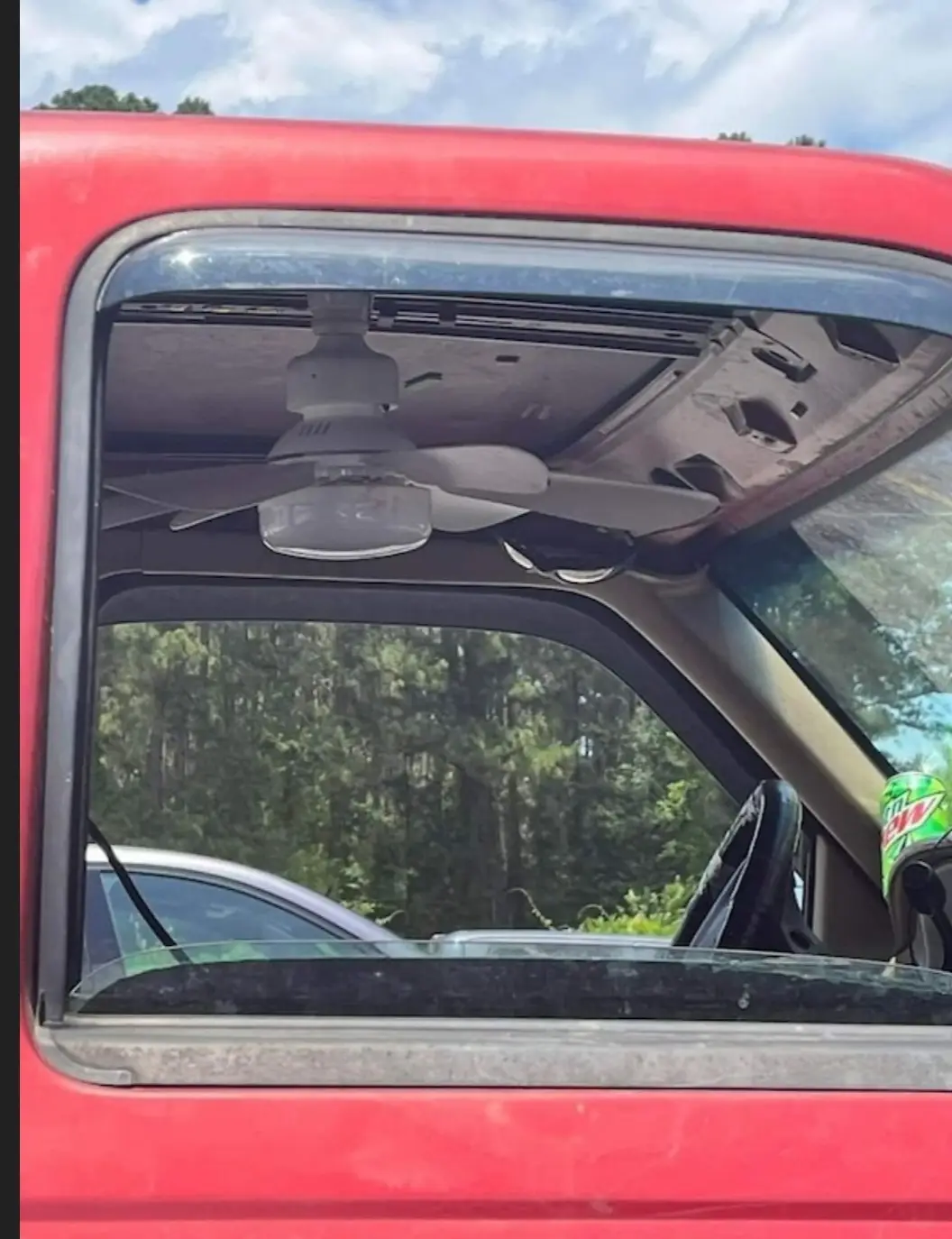 A white ceiling fan is visibly mounted to the interior roof of a red pickup truck, viewed from outside through the open…