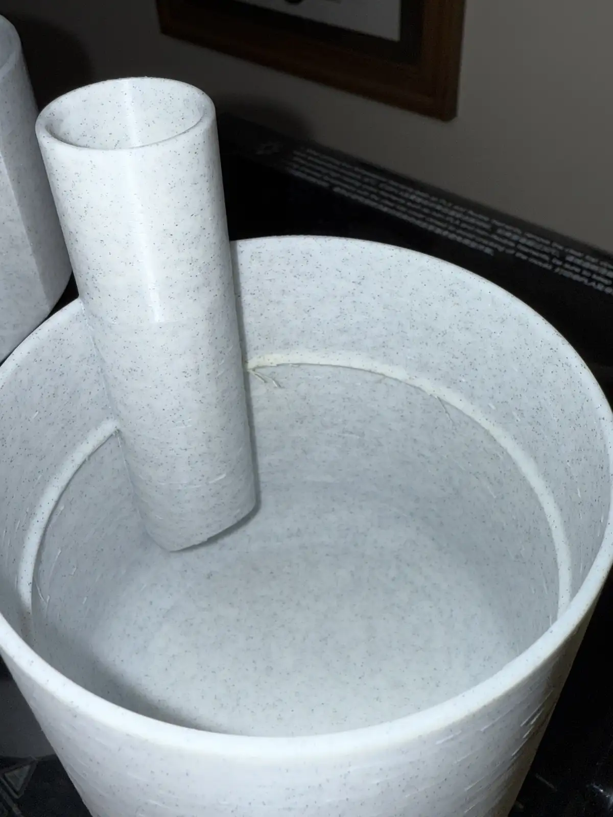 A close-up photo shows a white, speckled 3D-printed mortar and pestle resting on a dark surface, with visible layer lines…