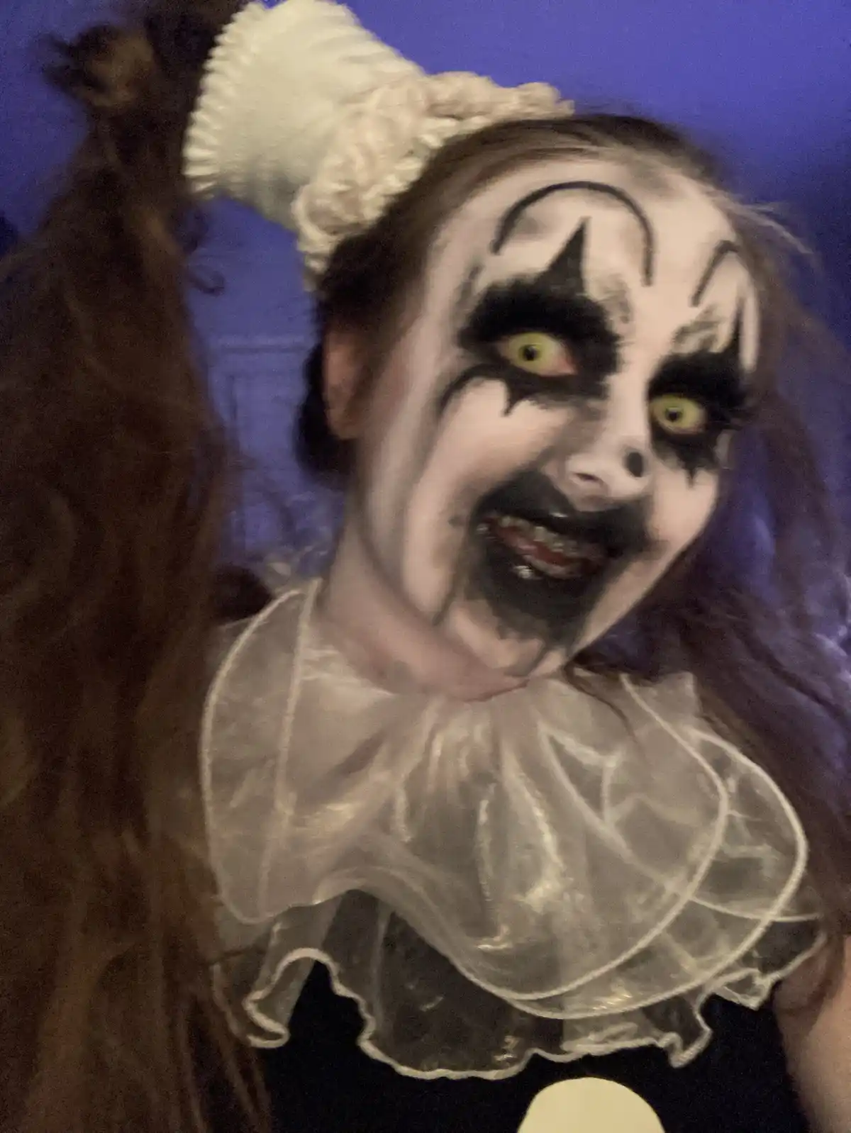 A person in a scary clown costume with white face paint, black makeup around eyes and mouth, yellow contact lenses, and a…