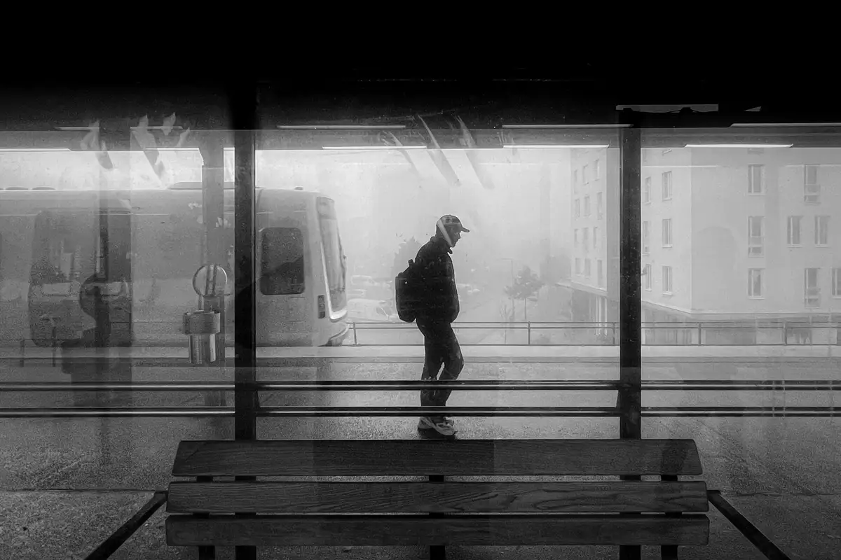 A black-and-white photo shows a person in silhouette walking through a bus or train station, viewed through glass panels…