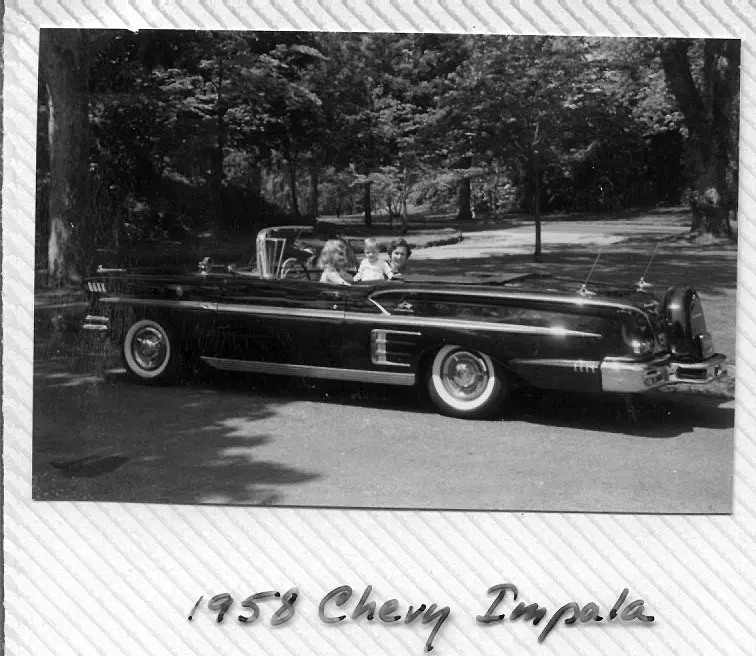 A vintage black-and-white photograph shows a 1958 Chevrolet Impala convertible parked on a tree-lined road, with three…