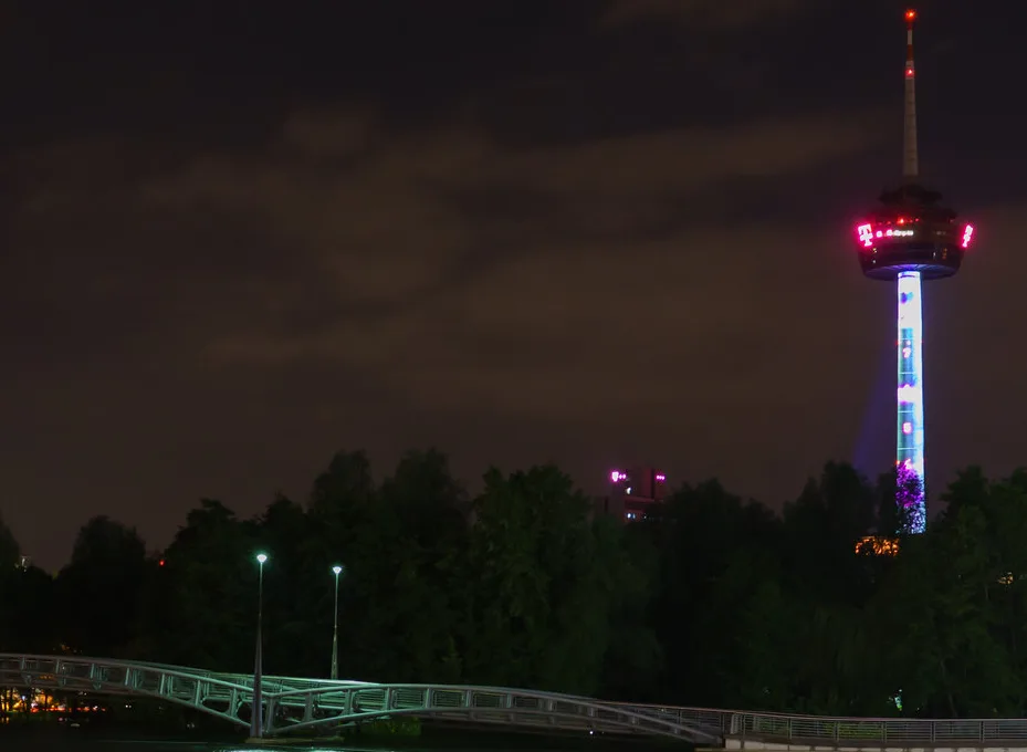 A nighttime photo of a tall, illuminated observation tower with red lights at the top and colorful vertical lights down its…