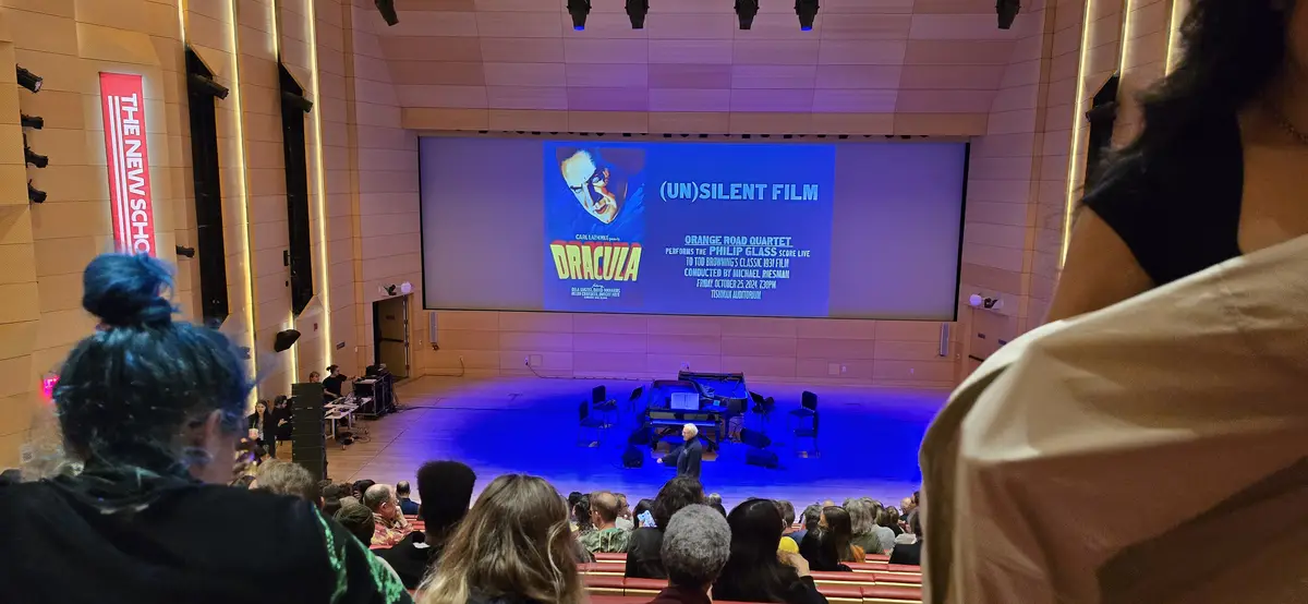 An audience watches a live performance in a theater where a large screen displays a poster for the 1931 film 'Dracula'…