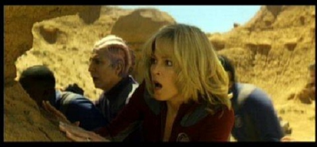 A still from a sci-fi movie shows a blonde woman in a red uniform with a shocked, wide-eyed expression, her mouth open in…