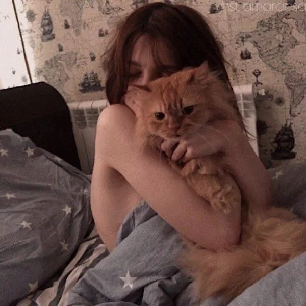 A person with long brown hair is lying in bed, hugging a fluffy orange cat tightly. The cat looks directly at the camera…