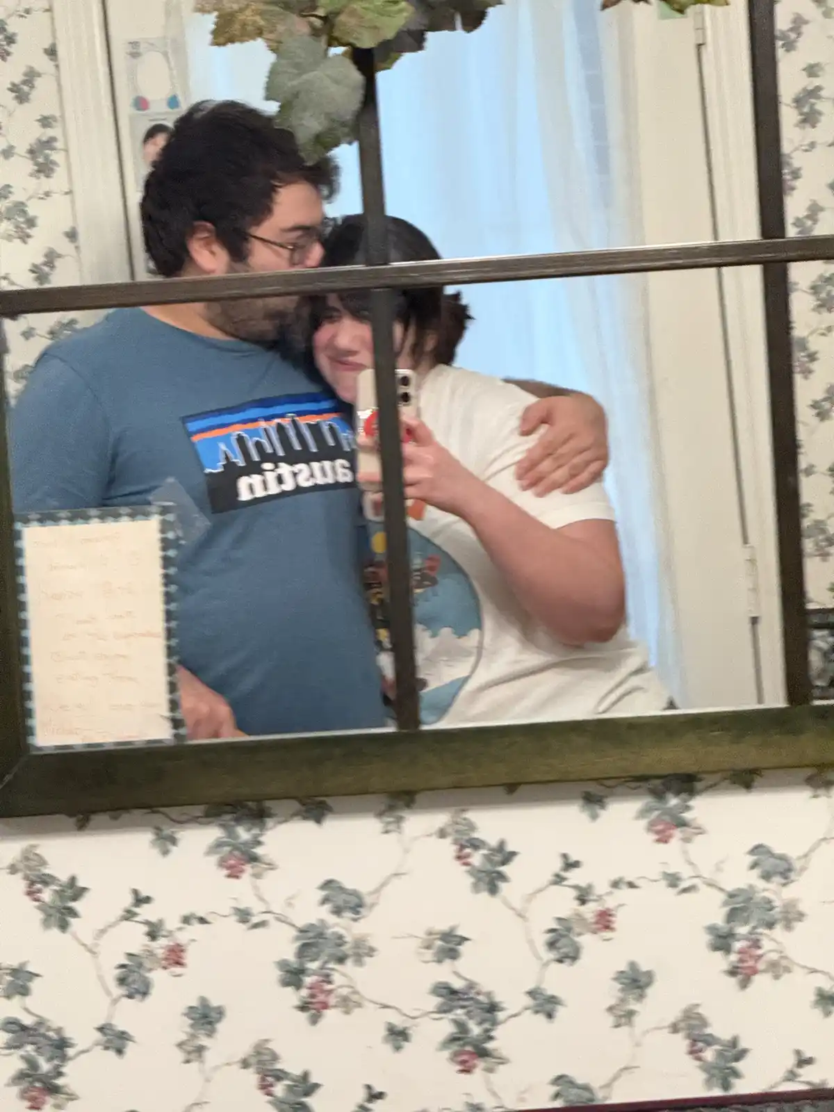A mirror selfie shows a man and woman embracing affectionately while the woman holds the phone. The man wears a blue…