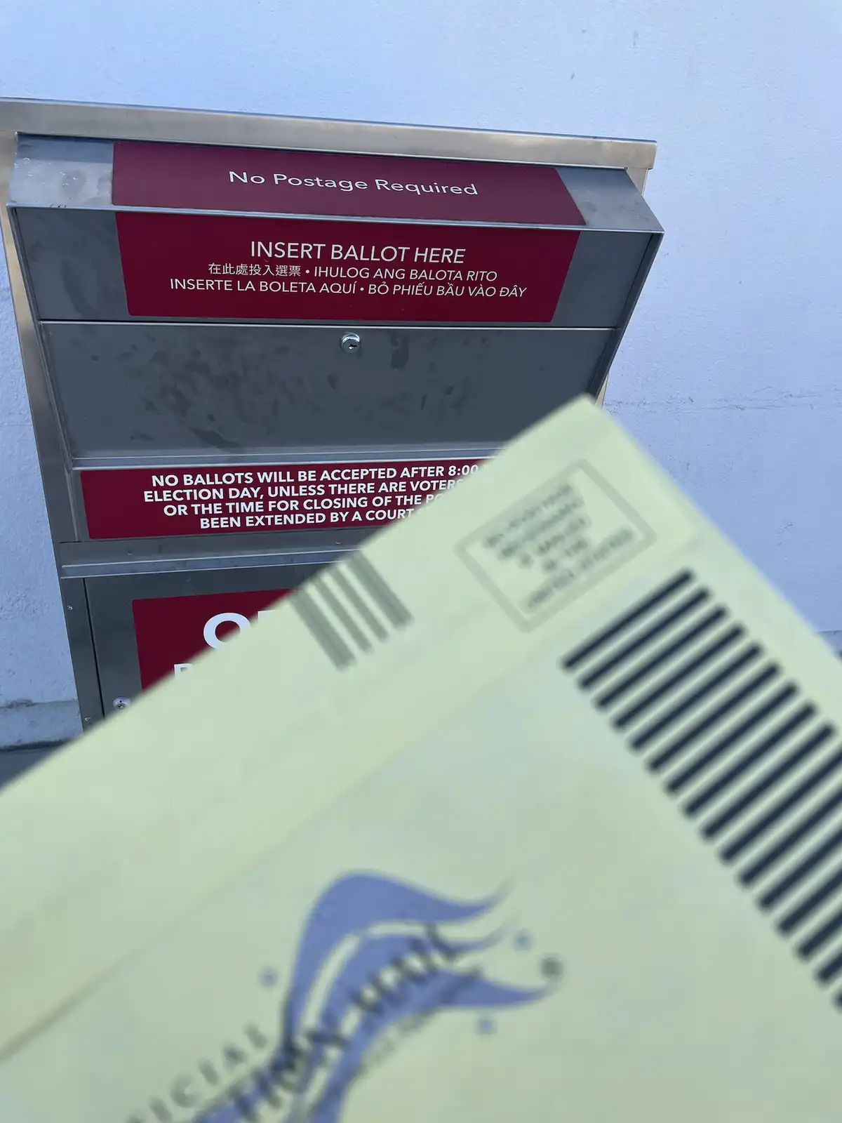 A close-up photo shows a light-colored ballot envelope in the foreground, partially obscuring a stainless steel ballot drop…