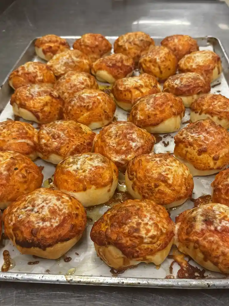 A close-up photo shows a metal baking tray filled with dozens of small, round, golden-brown baked goods topped with melted…