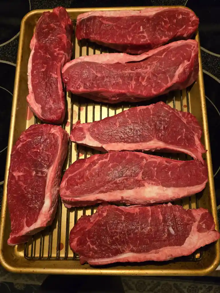 Seven raw, red beef steaks with visible marbling are arranged on a gold-colored metal baking tray with a wire rack. The…